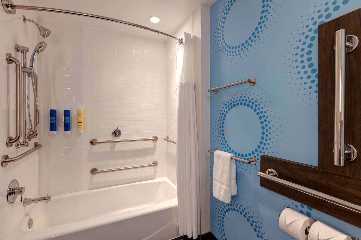 A modern bathroom with a white bathtub, shower, and handrails. There are toiletries on the wall and a towel rack.