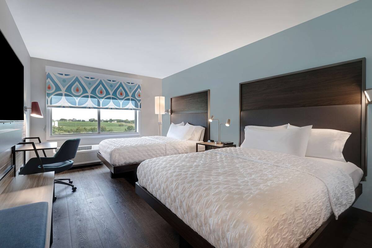 A hotel room with two beds, a desk, a chair, and a large window with a view of the outdoors.