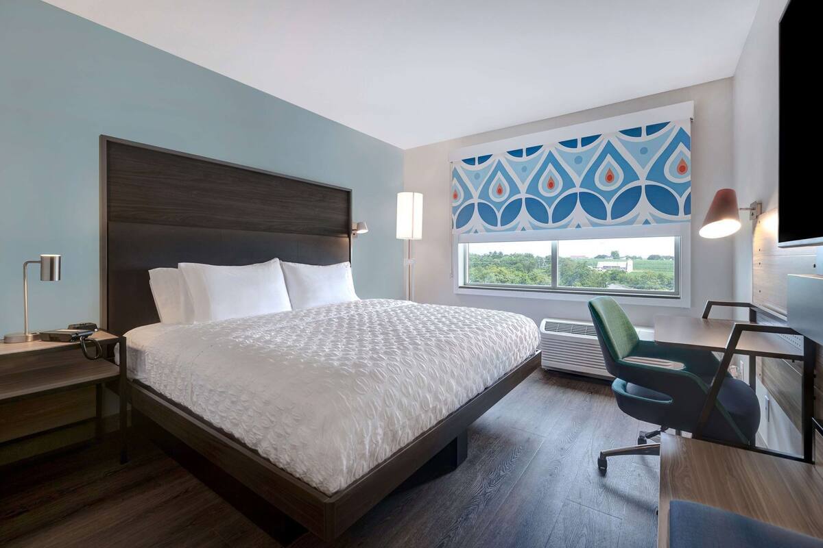 A modern hotel room with a large bed, a desk, a chair, a TV, and a window with a view.