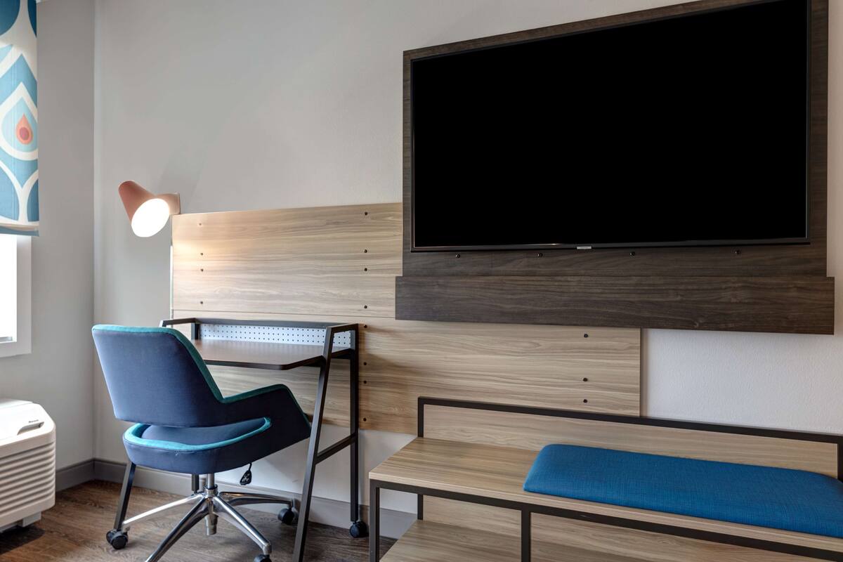 A modern room with a large flat-screen TV mounted on the wall, a desk with a chair, a bench, and a floor lamp.