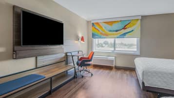 A hotel room with a bed, a desk with a chair, a TV mounted on the wall, a window with a view, and a painting on the wall.