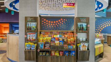 Un puesto de aperitivos con diversas opciones de comida empaquetada y un letrero que indica que es un mercado abierto las 24 horas.