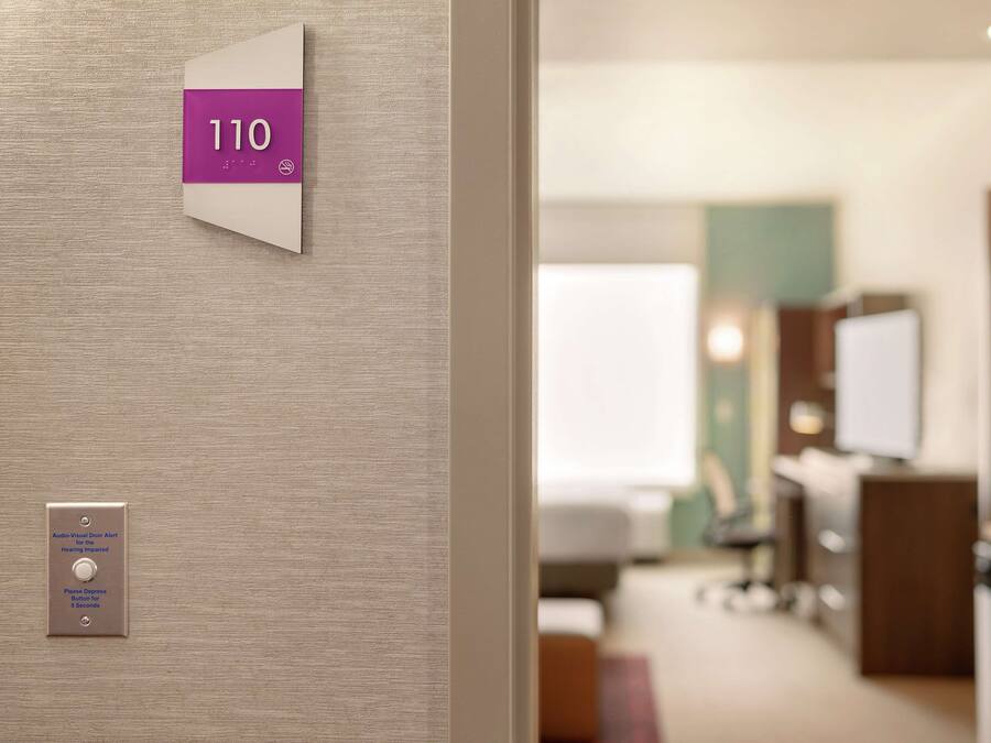 A hotel room with a number sign, an audio-visual door alarm, and a view of the room's interior including a bed, desk, and television.