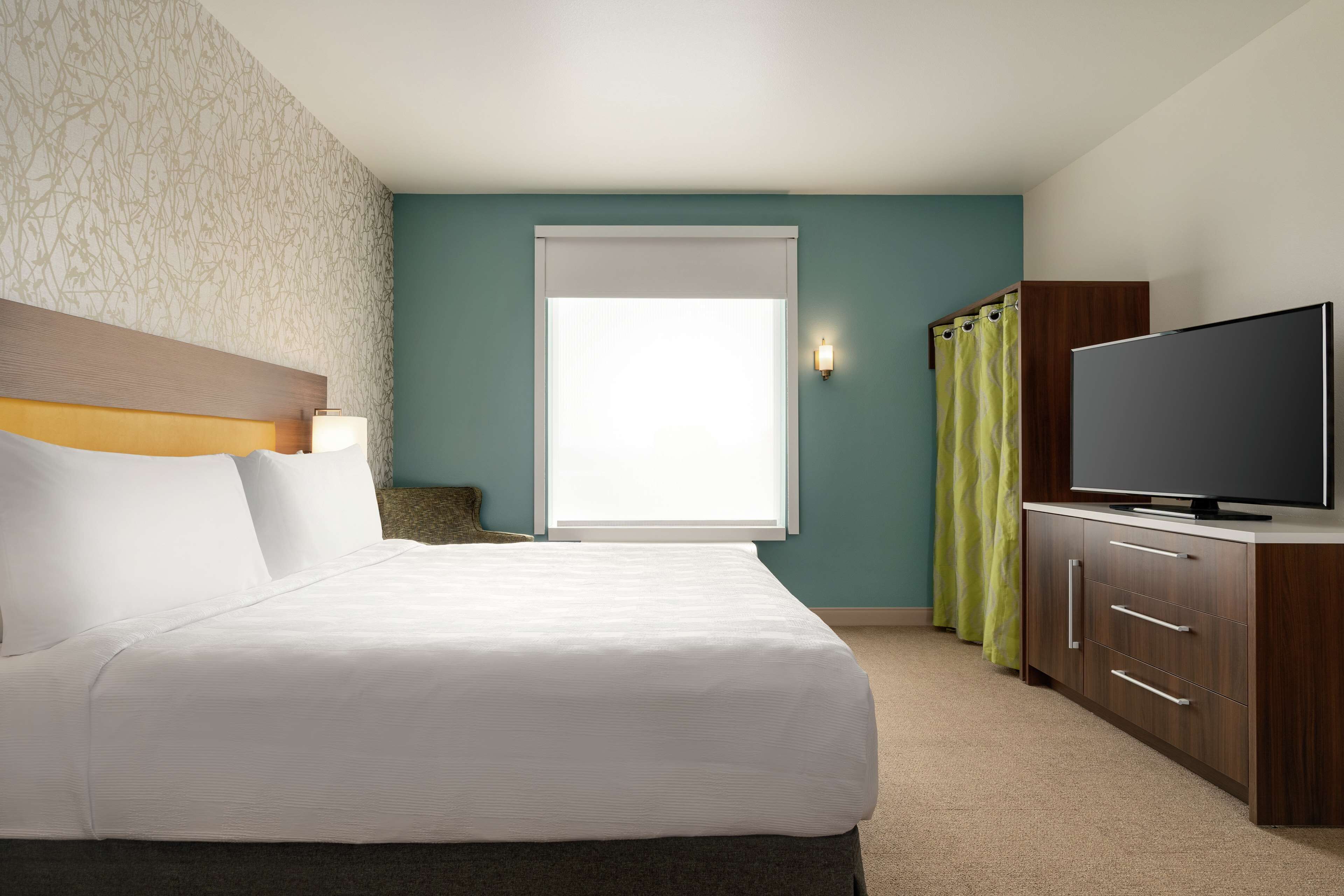 A hotel room with a bed, a television, a window with curtains, and a wall with a patterned design.