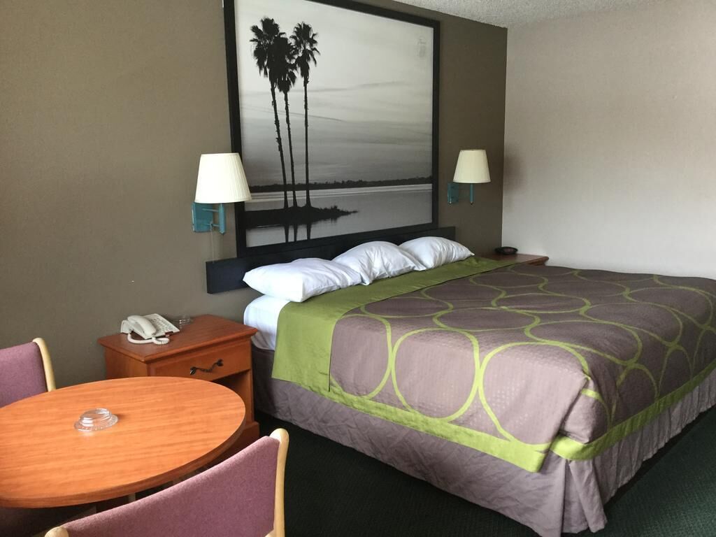 A hotel room with a bed, two bedside lamps, a nightstand with a phone, a round wooden table, and a painting of palm trees on the wall.