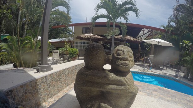 A stone statue near a poolside area with a tiled wall, palm trees, and a building with a red roof in the background.
