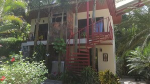 A two-story building with a red staircase and balconies, surrounded by lush greenery and palm trees.