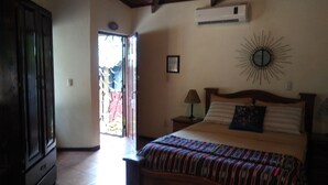 A bedroom with a bed, a nightstand, a lamp, a wall clock, and a door leading outside.