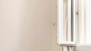 Una habitación en esquina con un sillón color beige, una ventana con cortinas y un equipo de aire acondicionado a la vista.