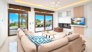 A modern living room with a large sofa, a coffee table, and a TV mounted on the wall. There are sliding glass doors leading to a balcony with outdoor furniture. - Sunlight Villa Heated Pool Sea View (Georgioupoli)