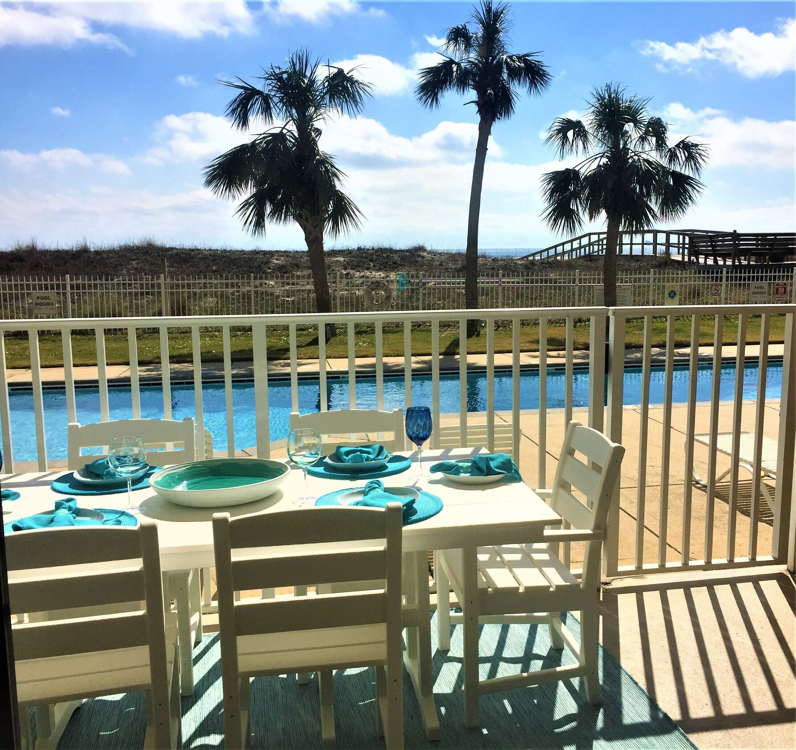A poolside dining area with white tables and chairs, a pool, and palm trees in the background.