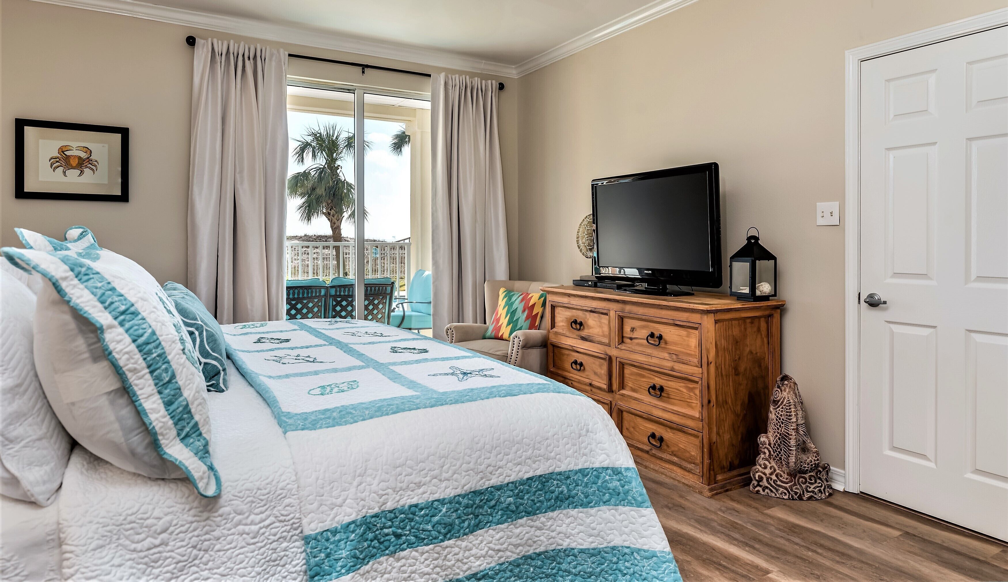 Un dormitorio con una cama, un televisor sobre un aparador de madera, una puerta y vistas a palmeras a través del ventanal.