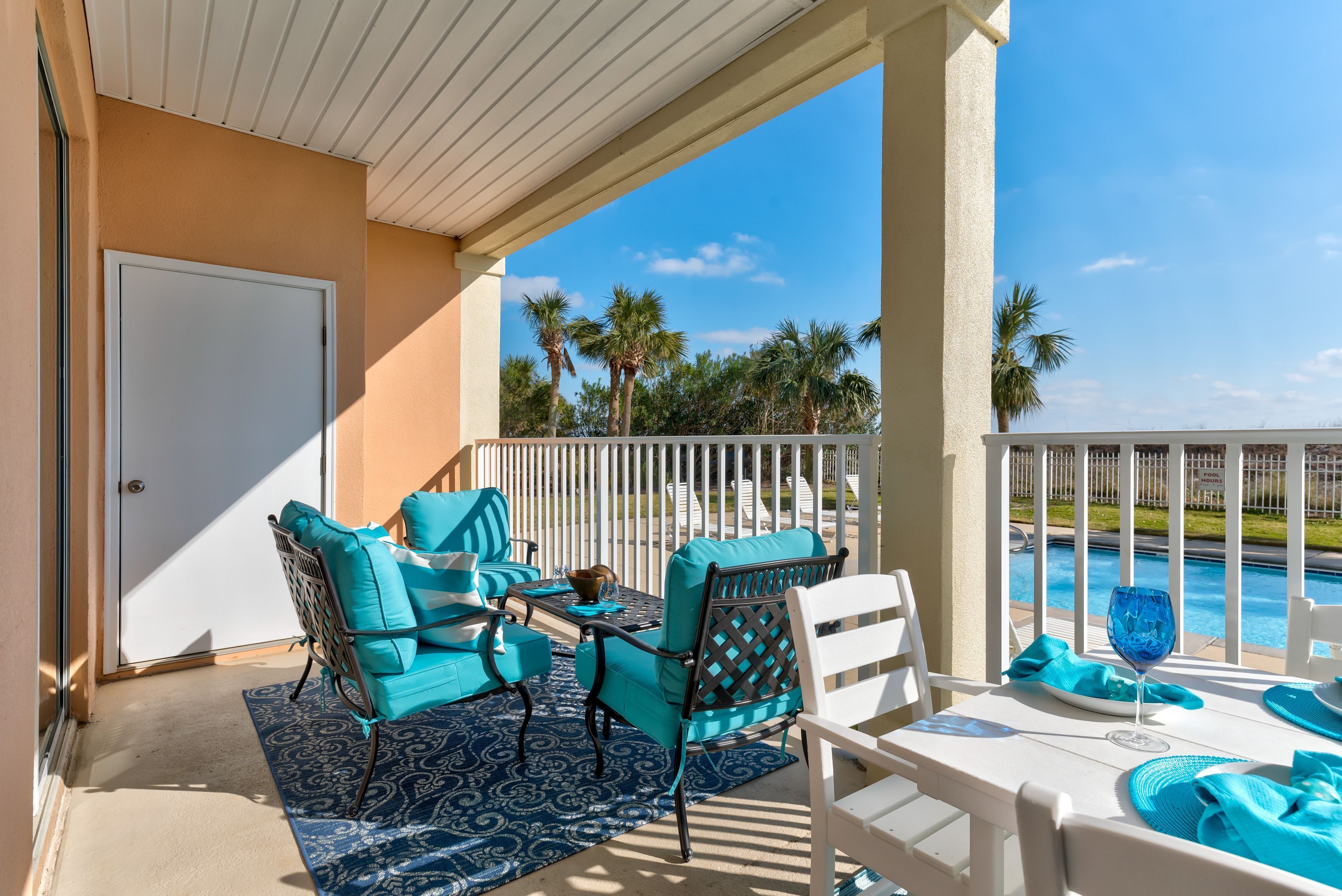 A covered outdoor seating area with turquoise chairs and a table, overlooking a pool and palm trees.