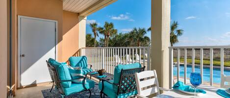 A covered outdoor seating area with turquoise chairs and a table, overlooking a pool and palm trees.