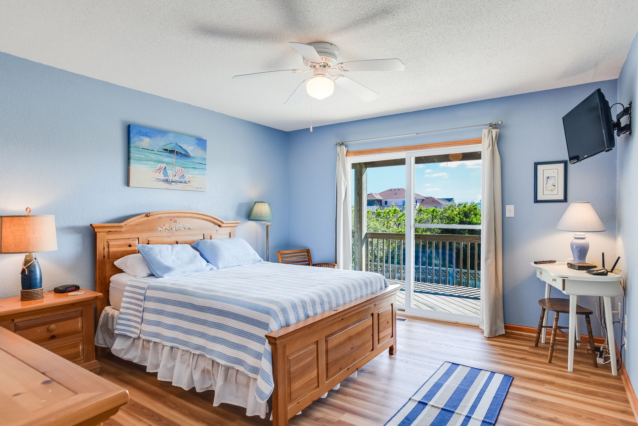 A bedroom with a bed, a desk, a chair, a ceiling fan, a TV, and a balcony with a view of houses and greenery.