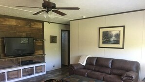A living room with a flat-screen TV on a wooden TV stand, a brown leather couch, a patterned rug, and a ceiling fan.