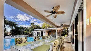 A poolside dining area with a glass table and chairs, a ceiling fan, and a view of the pool and surrounding greenery.
