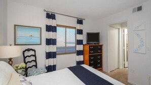 Quarto de hotel com uma cama, televisão sobre uma cômoda de madeira, um quadro na parede e vista para o mar pela janela.