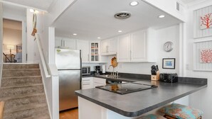 Uma cozinha moderna com armários brancos, uma geladeira de aço inoxidável, micro-ondas e fogão embutido. Há uma pequena ilha com banquetas e uma concha decorativa na parede.