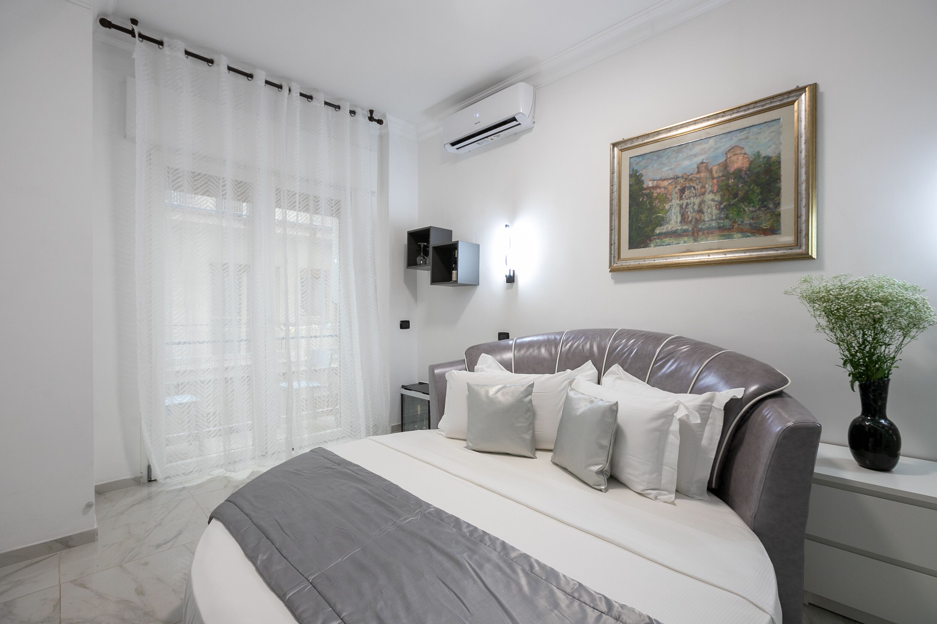 A modern bedroom with a large bed, a nightstand, a wall-mounted air conditioner, a framed painting, and a vase with flowers.