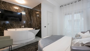 Une chambre d’hôtel moderne dotée d’une grande baignoire, d’un téléviseur à écran plat fixé au mur, d’un lit avec une couverture grise et d’une porte blanche.