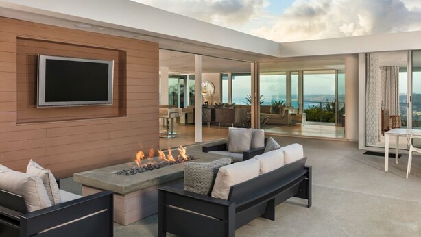 A modern outdoor living area with a fire feature, a flat-screen TV mounted on the wall, and a view of the cityscape through large glass doors. - Single Level Home with 2 Primary Bedrooms, Views and Spa in La Jolla Hills (La Jolla)