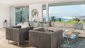A modern living room with a sofa, two armchairs, and a coffee table. There is a large window offering a view of the sea and greenery. - Single Level Home with 2 Primary Bedrooms, Views and Spa in La Jolla Hills (La Jolla)