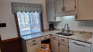 Una cocina compacta con armarios blancos, lavavajillas integrado, pileta doble y ventana con persianas.