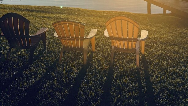 Una escena serena junto al lago, con dos sillas Adirondack mirando hacia un atardecer sobre el agua.