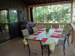 Un espace extérieur aménagé pour les repas, avec une table dressée pour quatre personnes, entourée de chaises, d’un barbecue et offrant une vue sur la forêt.