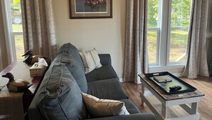 Uma sala de estar com um sofá seccional cinza, um quadro pendurado na parede e uma janela com cortinas.