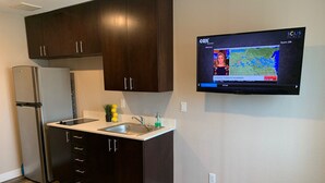 Una cocina moderna con armarios oscuros, un refrigerador de acero inoxidable, un lavamanos doble y un televisor de pantalla plana en la pared mostrando un pronóstico del tiempo.