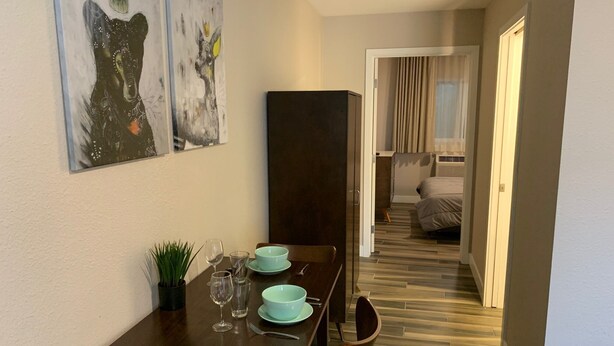 Un comedor con una mesa de madera, sillas y una planta pequeña. Hay dos cuadros en la pared y una puerta que da a un dormitorio.
