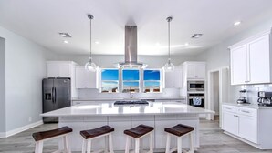 Une cuisine moderne dotée d’un îlot central, de meubles blancs, d’appareils électroménagers en acier inoxydable et offrant une vue sur le ciel grâce à de grandes fenêtres.