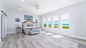 Une chambre avec un grand lit, un ventilateur de plafond et une vue sur la plage, visible à travers de nombreuses fenêtres.