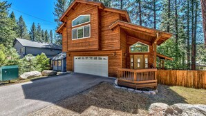 Une cabane en bois à deux étages, dotée d’un garage, entourée d’une terrasse en bois et d’une clôture, située dans une zone boisée.