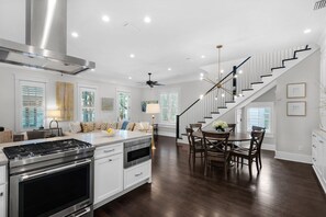A modern kitchen with a built-in stove, oven, and white cabinetry. There is a dining area with a wooden table and chairs, and a staircase with a black railing leading to an upper level. - Costello Cottage - Close to Cabana Pool & Butterfly Park! Free Fitness/Tennis! (Rosemary Beach)