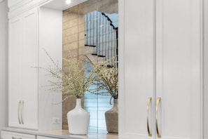 A white cabinet with gold handles, a white vase with branches, and a tiled wall with a staircase.