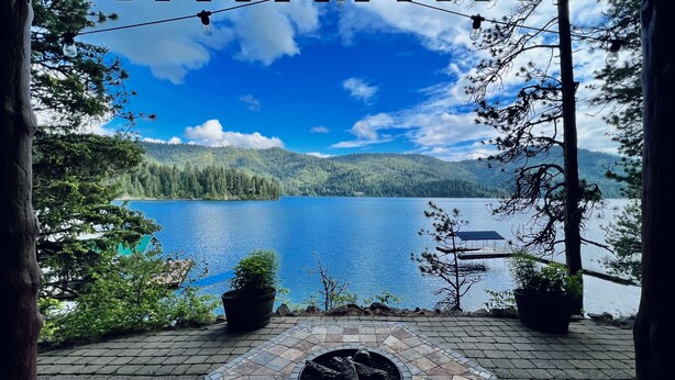 A patio with a stone fire pit, potted plants, and a view of a lake surrounded by trees and hills. - Pine View Cabin: Rustic Hayden Lake Cabin with Bunkhouse and Private Dock (Hayden)