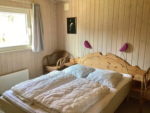 Un dormitorio con cabecera de madera, cama con ropa blanca, una silla de mimbre, una mesita de noche con una lámpara y una ventana con cortinas.