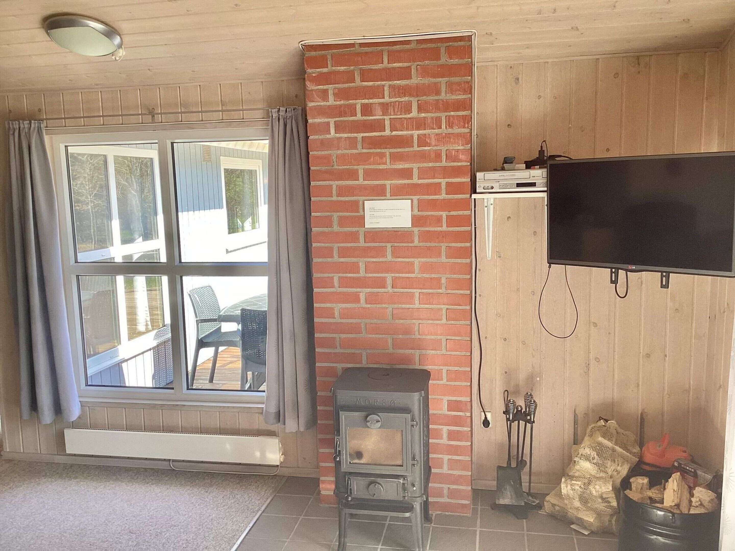 A room with a large window, a brick wall, a television mounted on the wall, and a wood-burning stove.