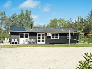 A small, single-story building with a dark exterior and a metal roof. There are two windows on the front side, a door, and a small porch area with four chairs. - Luxurious Holiday Home in Henne With Fireplace in the Garden (Henne)
