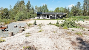 Une cabane moderne à deux étages dotée d’une grande terrasse, entourée d’un paysage naturel avec des arbres et une zone recouverte de gravier.