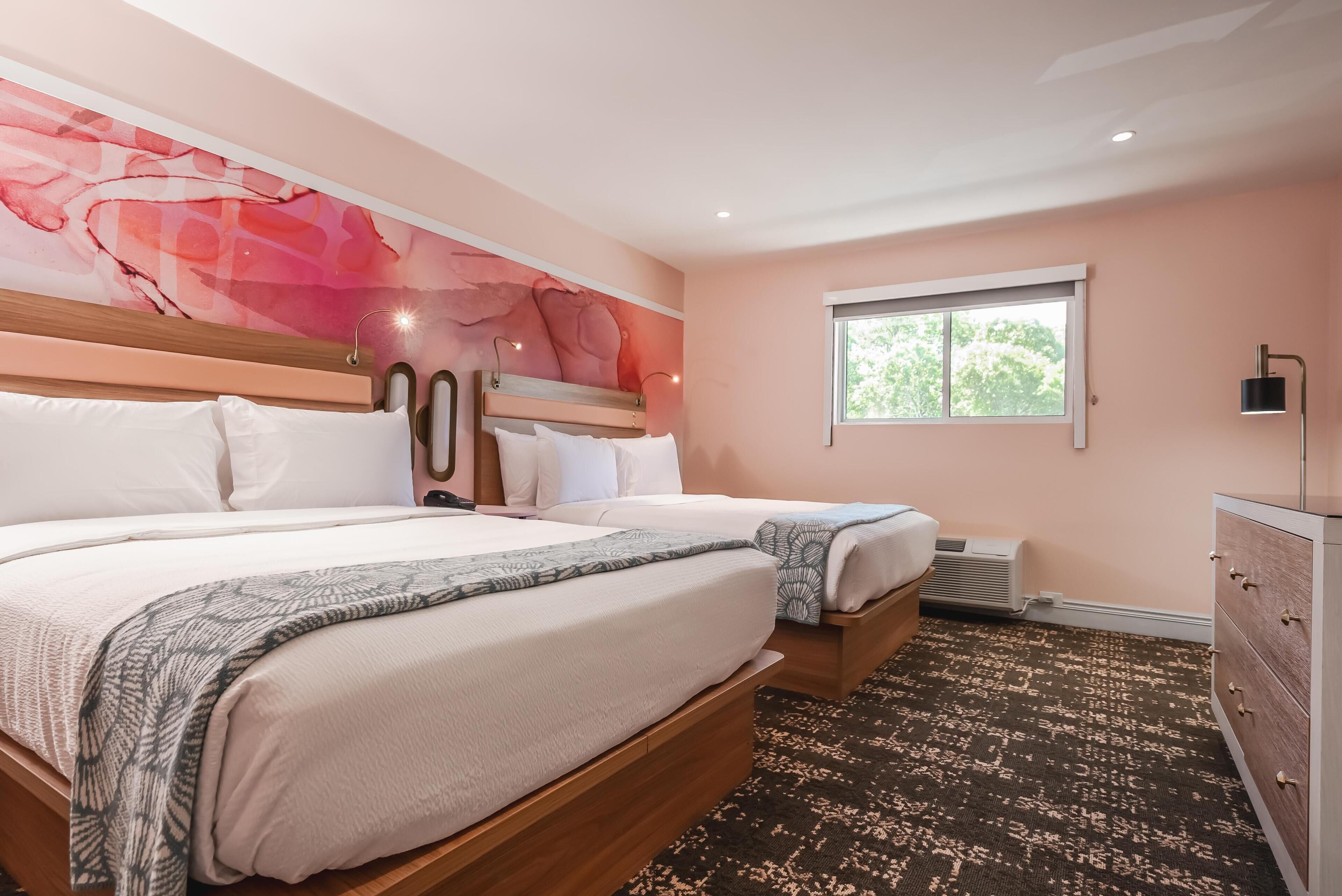 A hotel room with two beds, a large abstract mural on the wall, a window with a view of trees, and a dresser with drawers.