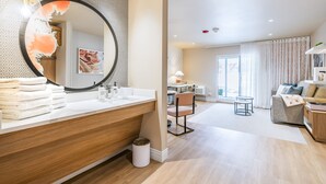 A modern bathroom with a large mirror, wooden vanity, and a sink. There is a chair and a small table in the adjacent living area, with a sofa and a view of the outdoors. - The Bentley Hotel Southampton (Southampton)