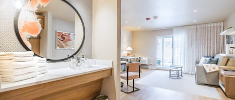 Ein modernes Badezimmer mit einem großen Spiegel, einem Holzausleger und einem Waschbecken. Im angrenzenden Wohnbereich befinden sich ein Sessel und ein kleiner Tisch, ein Sofa und ein Blick ins Freie.