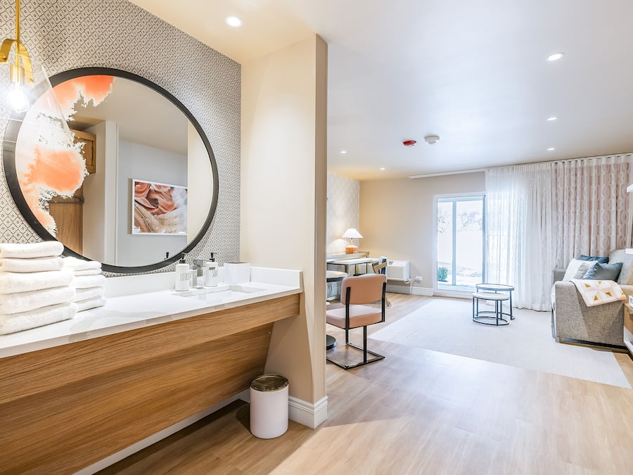A modern bathroom with a large mirror, wooden vanity, and a sink. There is a chair and a small table in the adjacent living area, with a sofa and a view of the outdoors.
