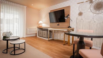 Une chambre d’hôtel moderne avec un bureau en bois, un téléviseur à écran plat, une table ronde en verre et un coin salon agrémenté d’un fauteuil rose.