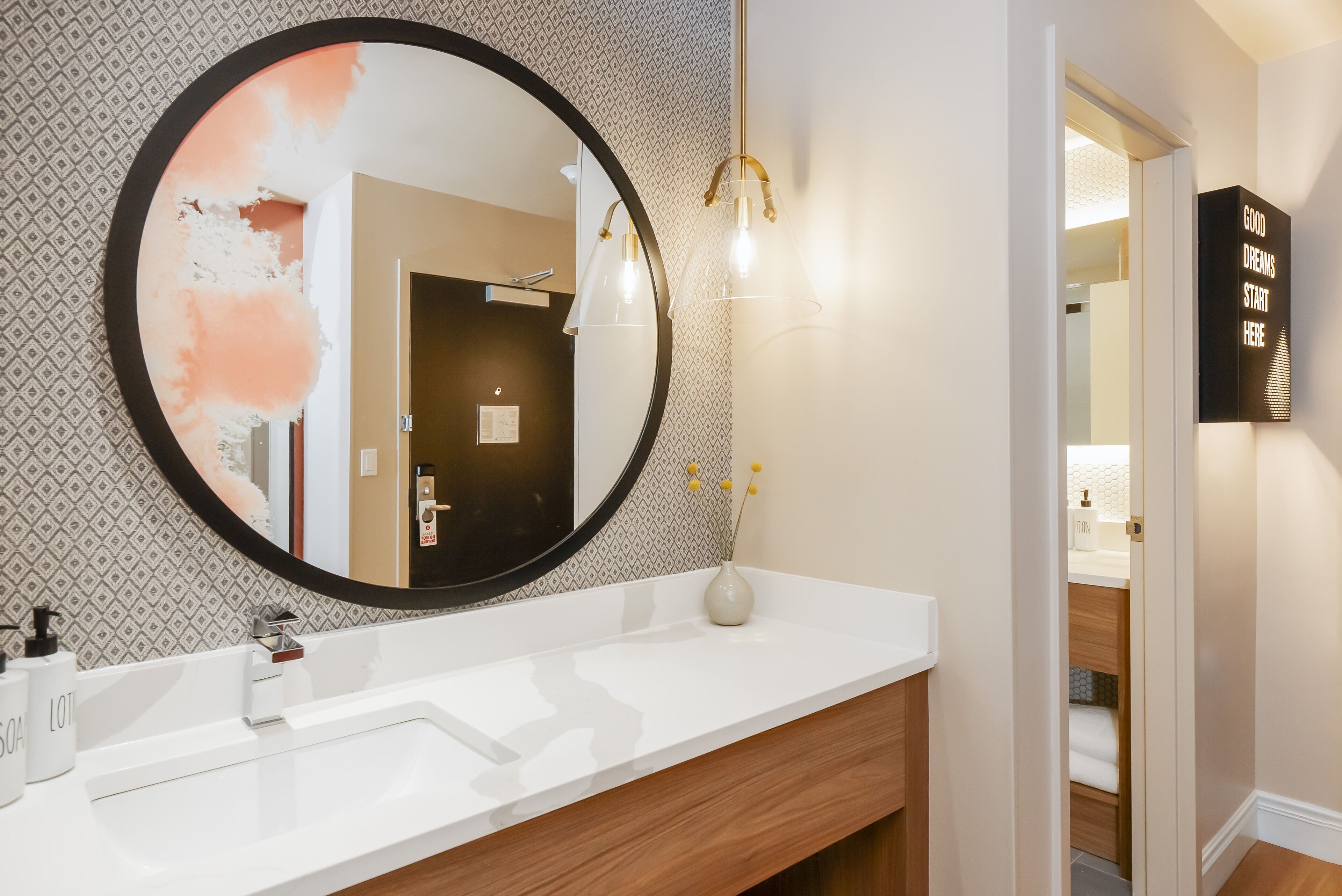Une salle de bain avec un miroir rond, un lavabo blanc et un meuble-vasque en bois. Un luminaire est fixé au mur au-dessus du miroir.