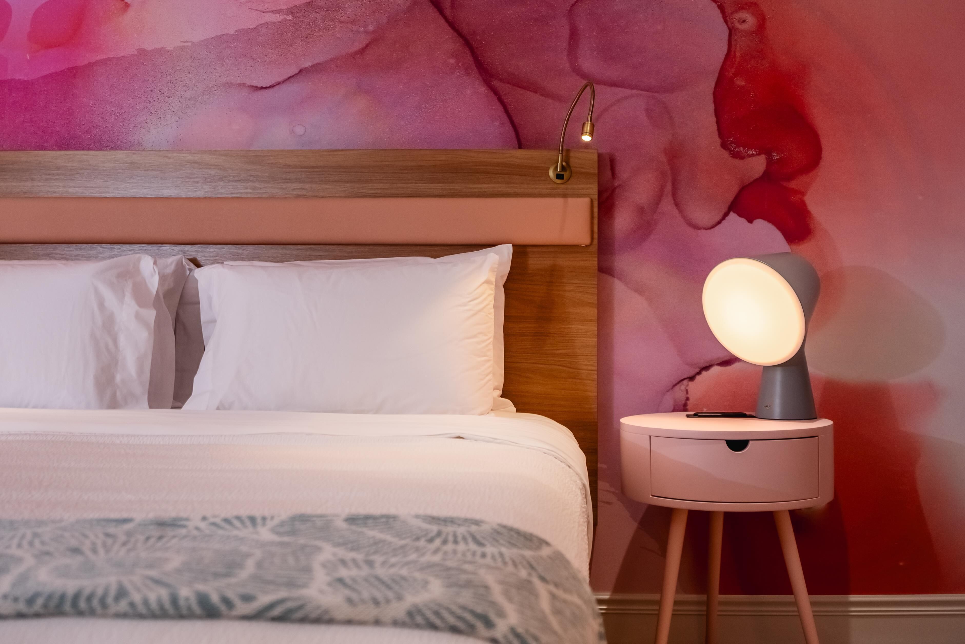 A neatly made bed with white linens and a patterned throw blanket, a bedside table with a lamp, and a vibrant abstract wall mural.